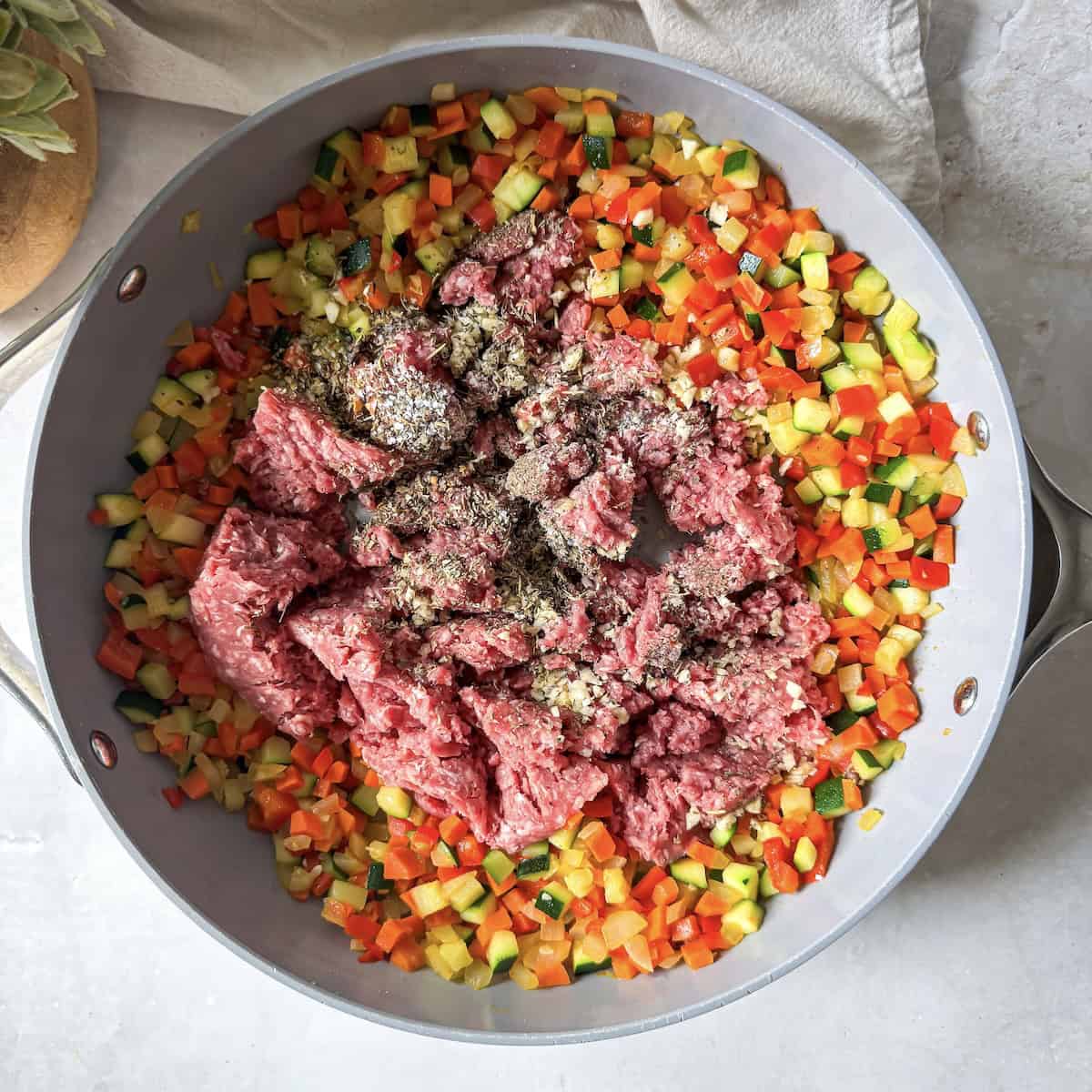 Ground beef and seasonings added to cooked vegetables in a skillet.