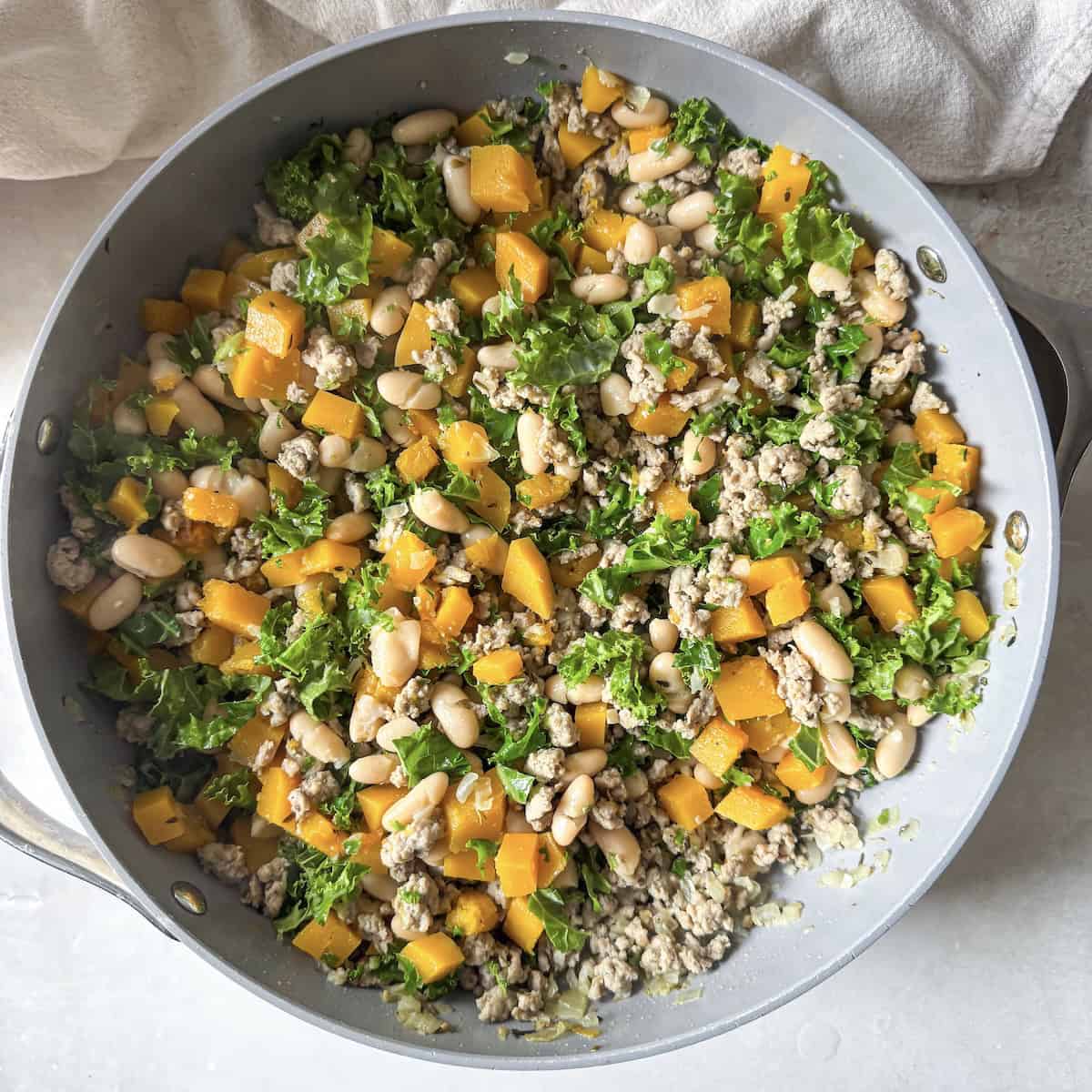 A skillet with cooked ground turkey, cubed butternut squash, kale, white beans, all stirred together just before the kale is cooked.