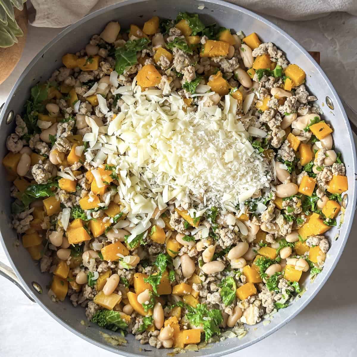 A skillet with ground turkey, kale, butternut squash, and white beans with mozzarella and parmesan cheese added to it just before the cheese is stirred in and melted.