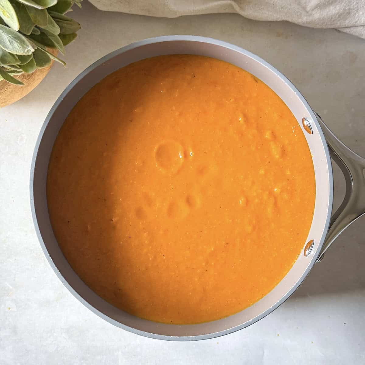 a bright orange smoothly blended soup in a big pot on a gray and white background.
