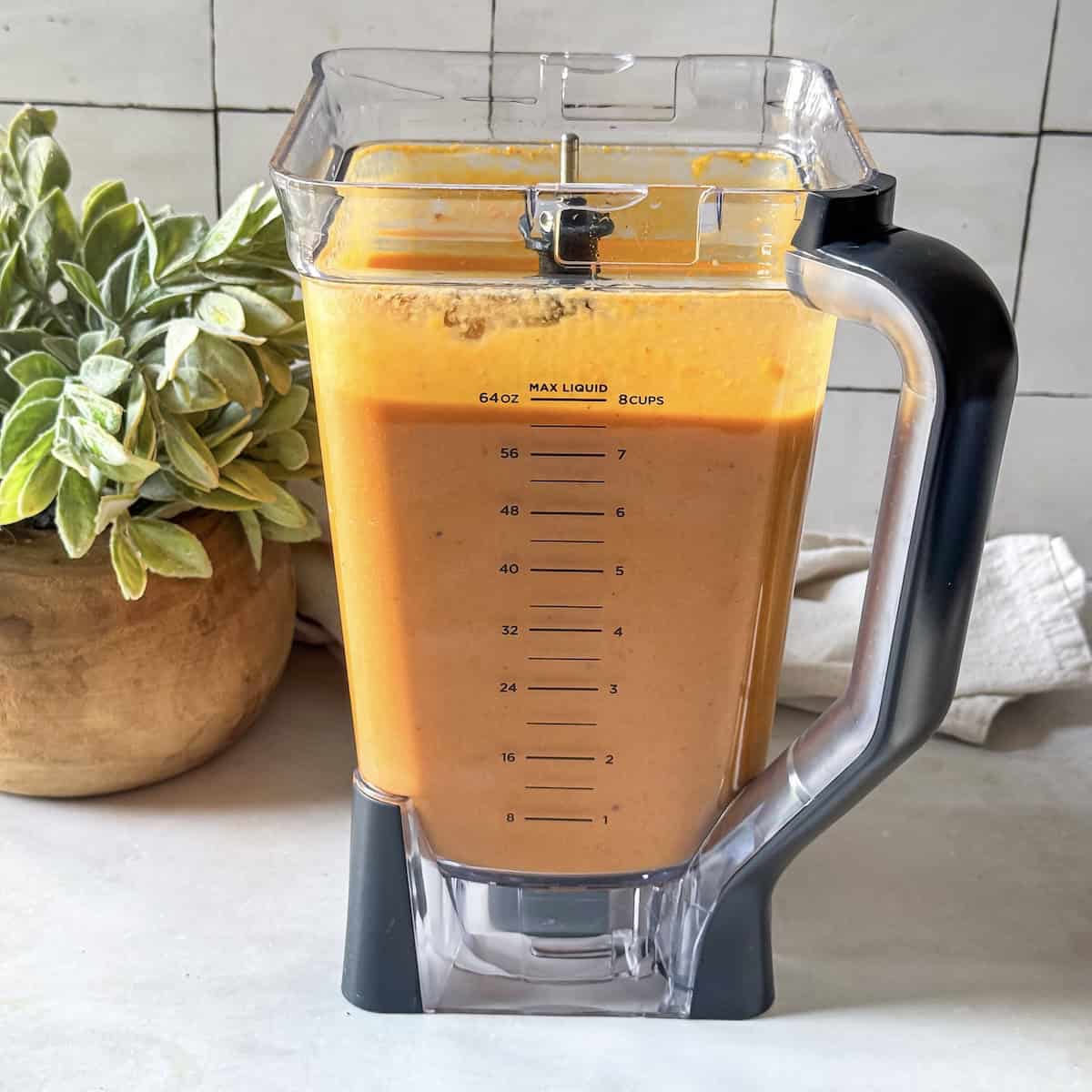 a bright orange soup in a blend right after it's been blended with a green plant in the background.