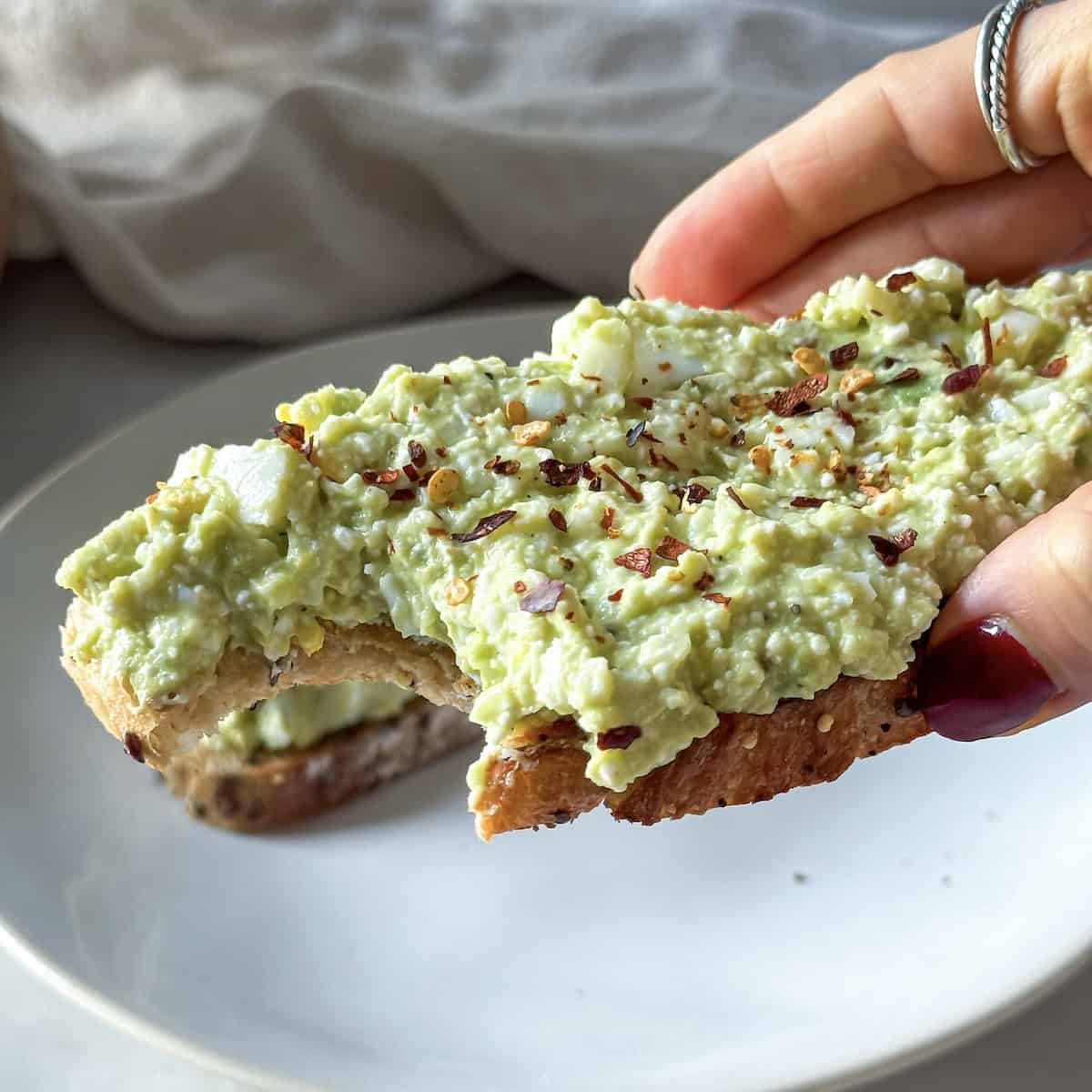a human hand holding a slice of avocado toast with cottage cheese and egg with a bite out of it.