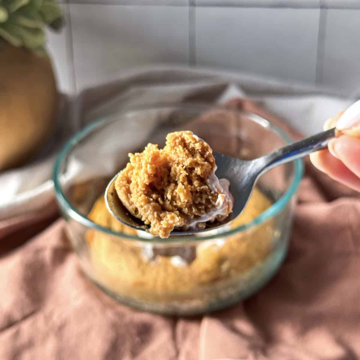 a human hand holding a spoon with a bite of fluffy pumpkin protein pancake bowl on it and the rest of the pancake bowl in the background in a glass bowl.