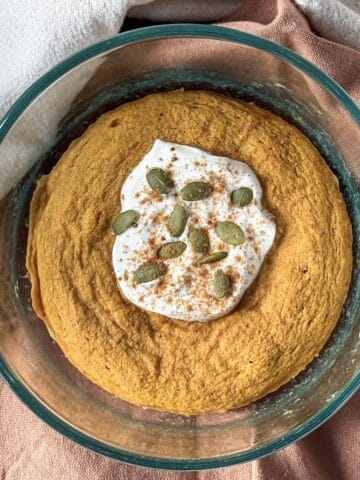 a pumpkin protein pancake bowl after being cooked in a glass bowl with a scoop of greek yogurt, pumpkin seeds, and pumpkin pie spice on top on a burnt orange and tan dishcloth.