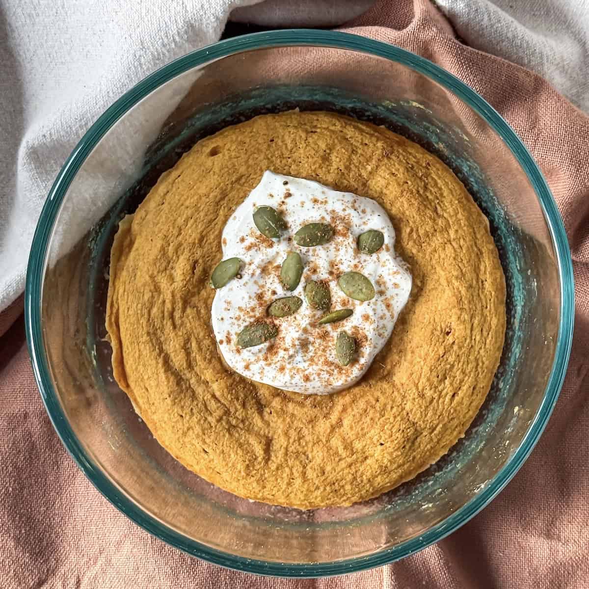 a pumpkin protein pancake bowl after being cooked in a glass bowl with a scoop of greek yogurt, pumpkin seeds, and pumpkin pie spice on top on a burnt orange and tan dishcloth.
