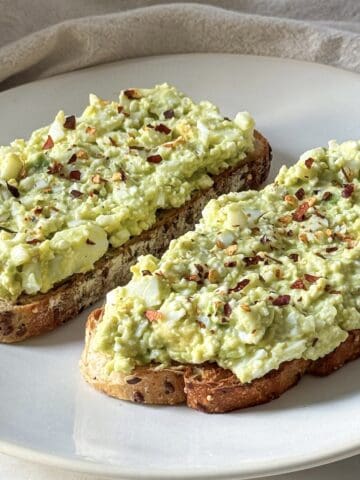 2 slices of avocado toast with cottage cheese and egg on a white plate with red pepper flakes on top.
