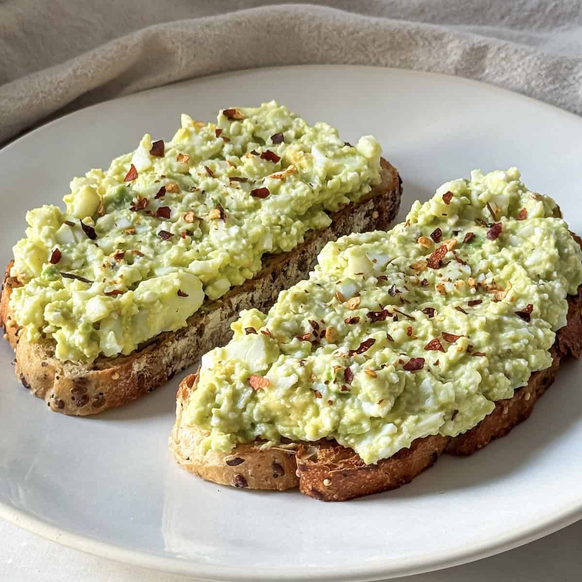 2 slices of avocado toast with cottage cheese and egg on a white plate with red pepper flakes on top.