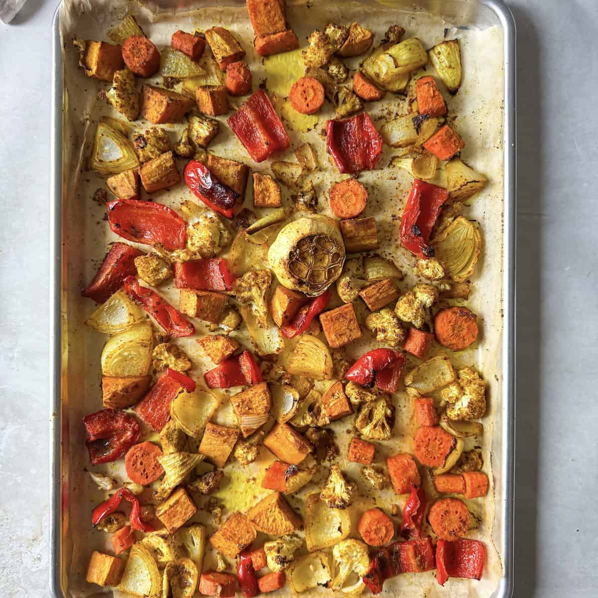 Colorful diced vegetables including bell pepper, carrots, sweet potatoes, cauliflower, onion, a bulb of garlic, and fresh ginger seasoned and on a sheet pan lined with parchment paper after they've been roasted.