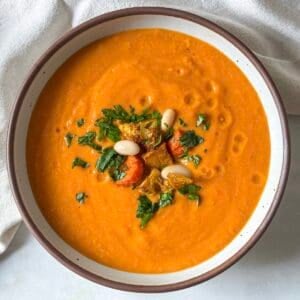 A golden soup in a big white bowl with brown speckles and a tan rim. The soup is garnished with chopped fresh parsley, white beans, and a couple pieces of roasted vegetables with a light tan dish towel in the background