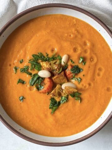 A golden soup in a big white bowl with brown speckles and a tan rim. The soup is garnished with chopped fresh parsley, white beans, and a couple pieces of roasted vegetables with a light tan dish towel in the background