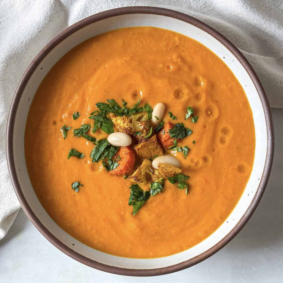 A golden soup in a big white bowl with brown speckles and a tan rim. The soup is garnished with chopped fresh parsley, white beans, and a couple pieces of roasted vegetables with a light tan dish towel in the background