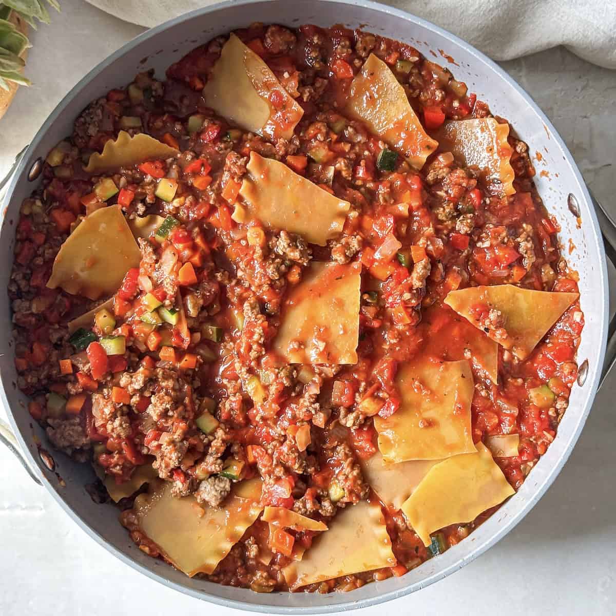 lasagna noodles added to crushed tomatoes and ground beef with cooked veggies in a skillet.