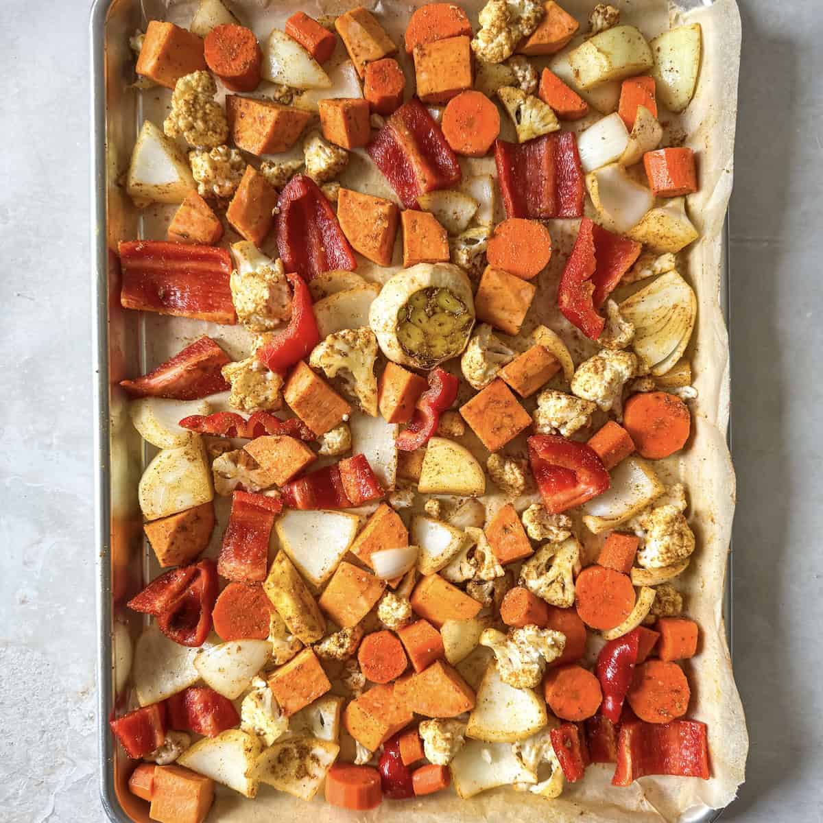 overhead view of diced vegetables including bell pepper, carrots, sweet potatoes, cauliflower, onion, a bulb of garlic, and fresh ginger seasoned and on a sheet pan lined with parchment paper before being roasted.