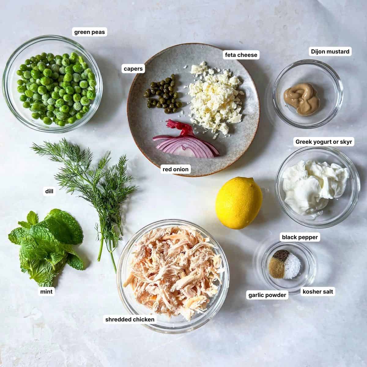 Ingredients for chicken salad with peas in separate containers on a table with a grayish white background labeled.