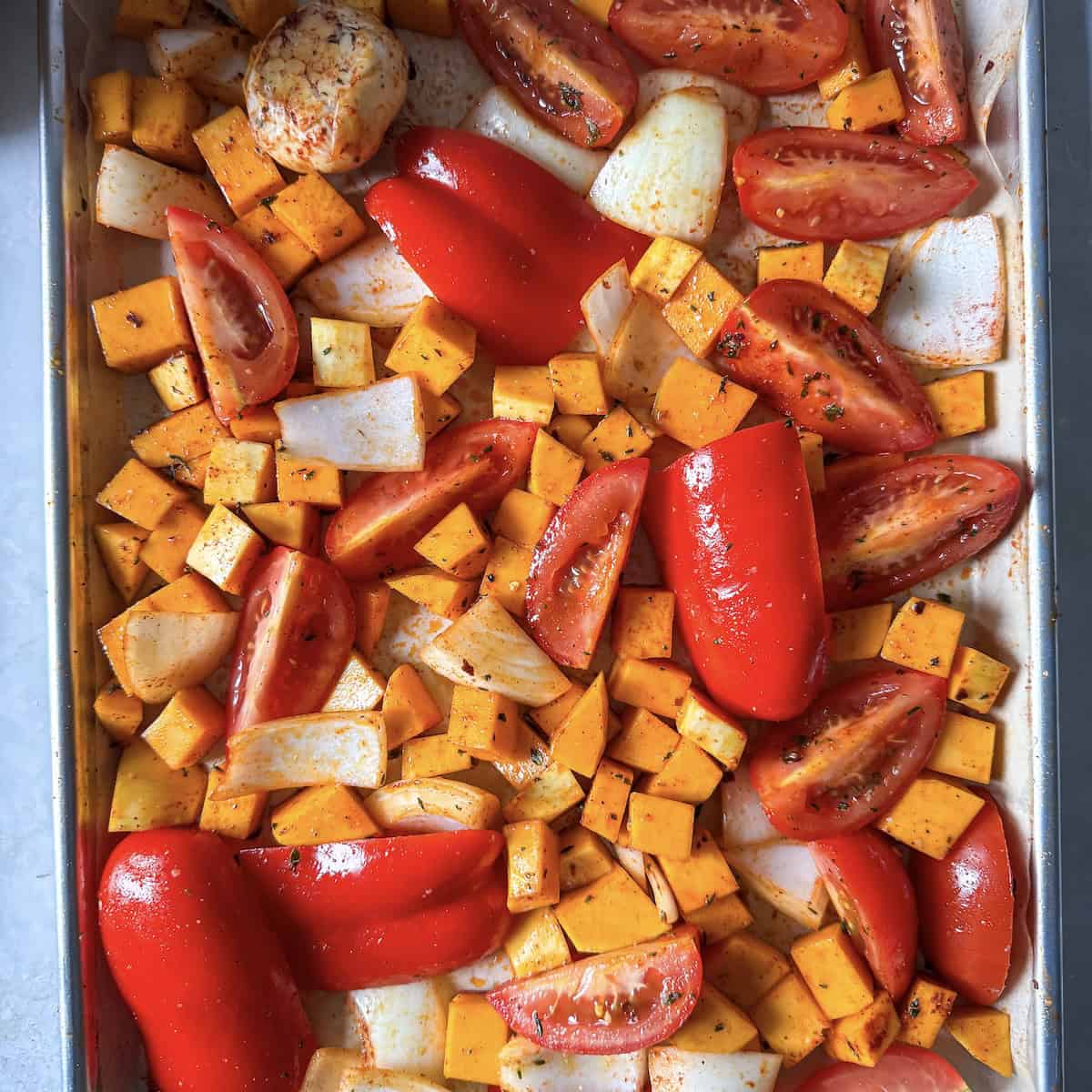 Diced butternut squash, quartered roma tomatoes, red bell pepper, yellow onion, and a bulb of garlic on a sheet pan after being tossed with olive oil and seasonings before going in the oven.