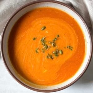 A big bowl of butternut squash tomato soup garnished wit hfresh thyme in a white bowl with a tan edge and speckles.