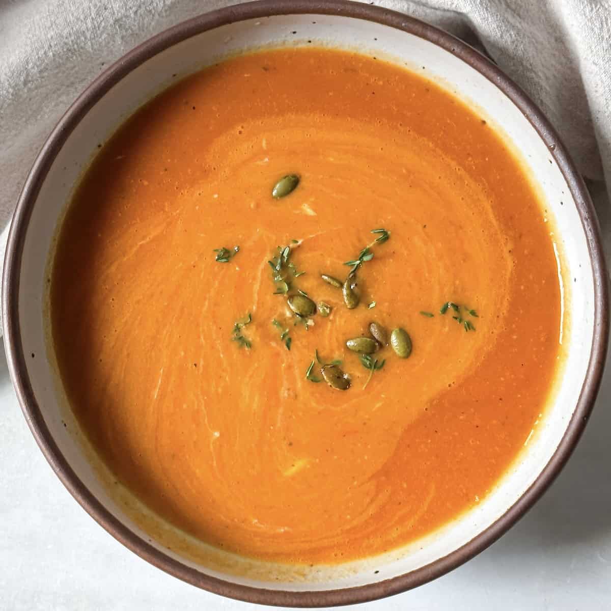 A big bowl of butternut squash tomato soup garnished wit hfresh thyme in a white bowl with a tan edge and speckles.