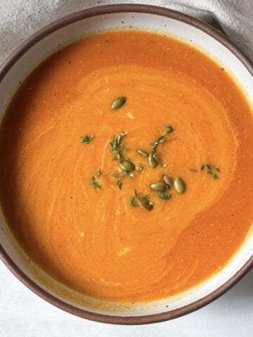 A big bowl of butternut squash tomato soup garnished wit hfresh thyme in a white bowl with a tan edge and speckles.